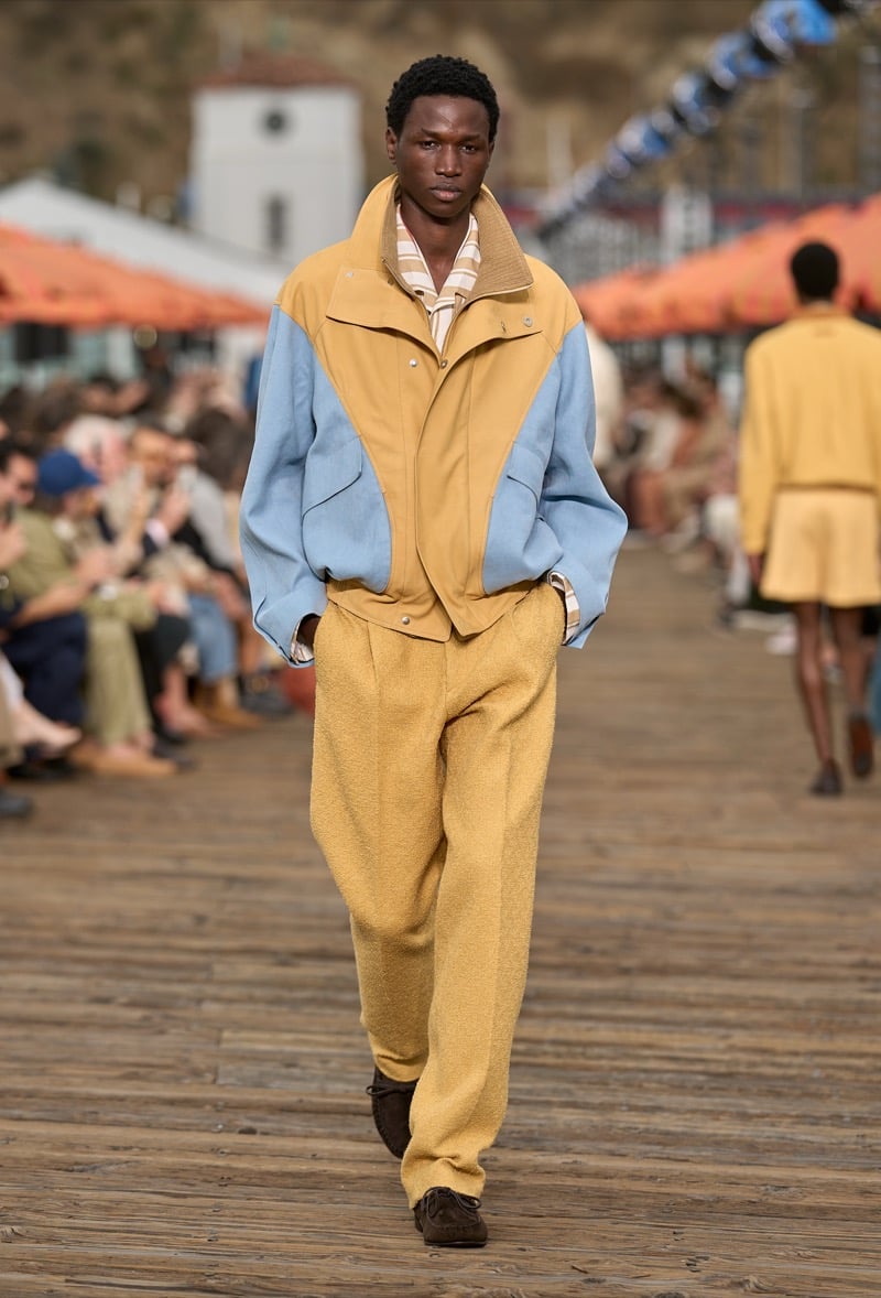 Male model wears a pale blue and butter yellow color-blocked windbreaker over yellow boucle wide-leg trousers with brown suede shoes