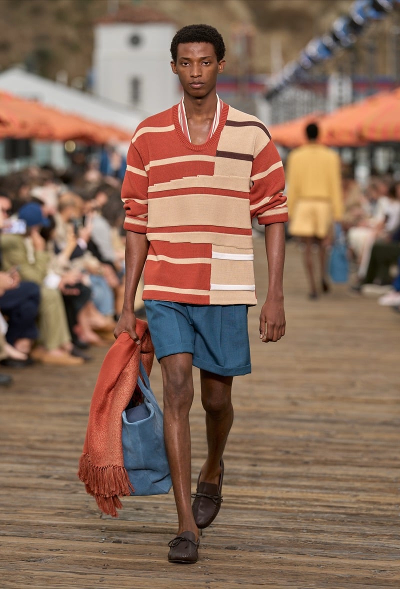 Male model wears an oversized rust, cream, and white patchwork striped knit pullover over teal blue pleated shorts with brown moccasins, carrying a blue tote with a rust throw