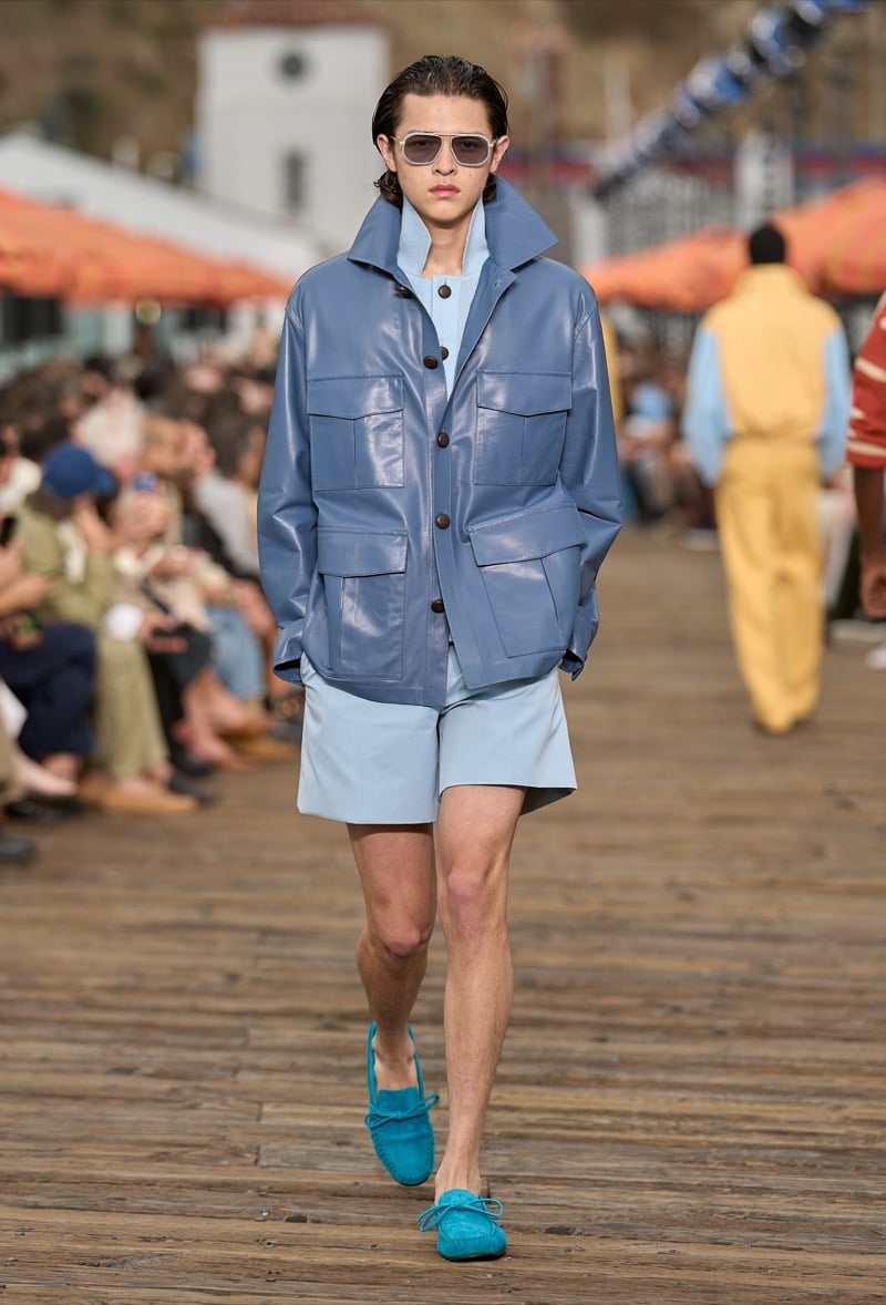 Male model wears a powder blue leather utility jacket with cargo pockets over a pale blue half-zip pullover and matching pale blue shorts with turquoise suede moccasins