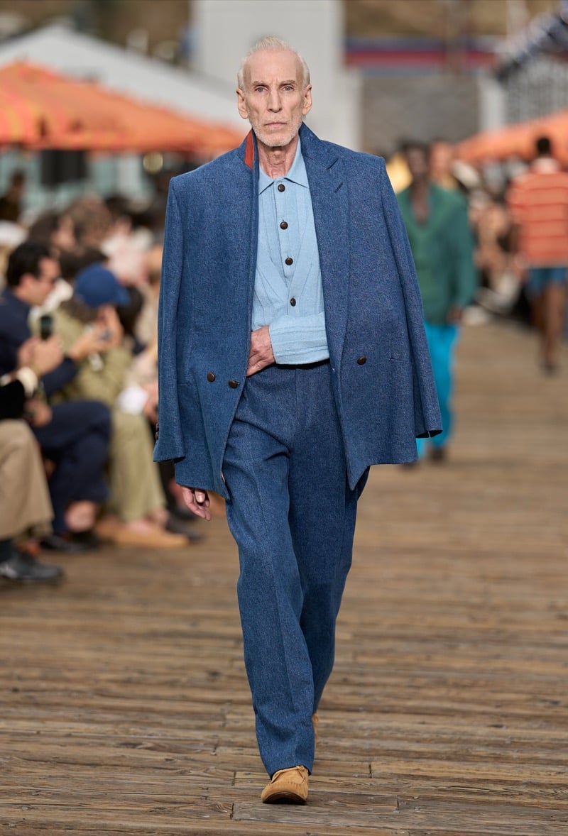 Male model wears a denim blue double-breasted blazer draped over the shoulders with a pale blue knit polo and matching denim blue wide-leg trousers
