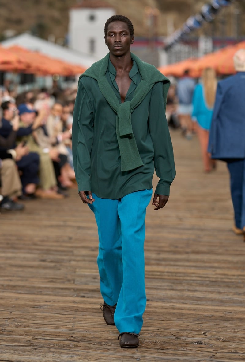 Male model wears a forest green open-collar silk shirt with a sage knit tied at the shoulders over bright turquoise wide-leg trousers and brown moccasins
