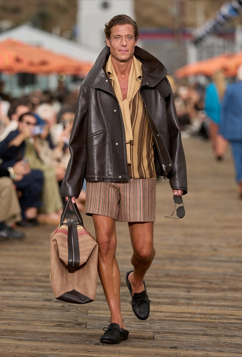 Male model wears an oversized dark brown leather bomber with funnel collar over a tan and rust striped silk shirt with matching striped shorts, holding a striped weekender
