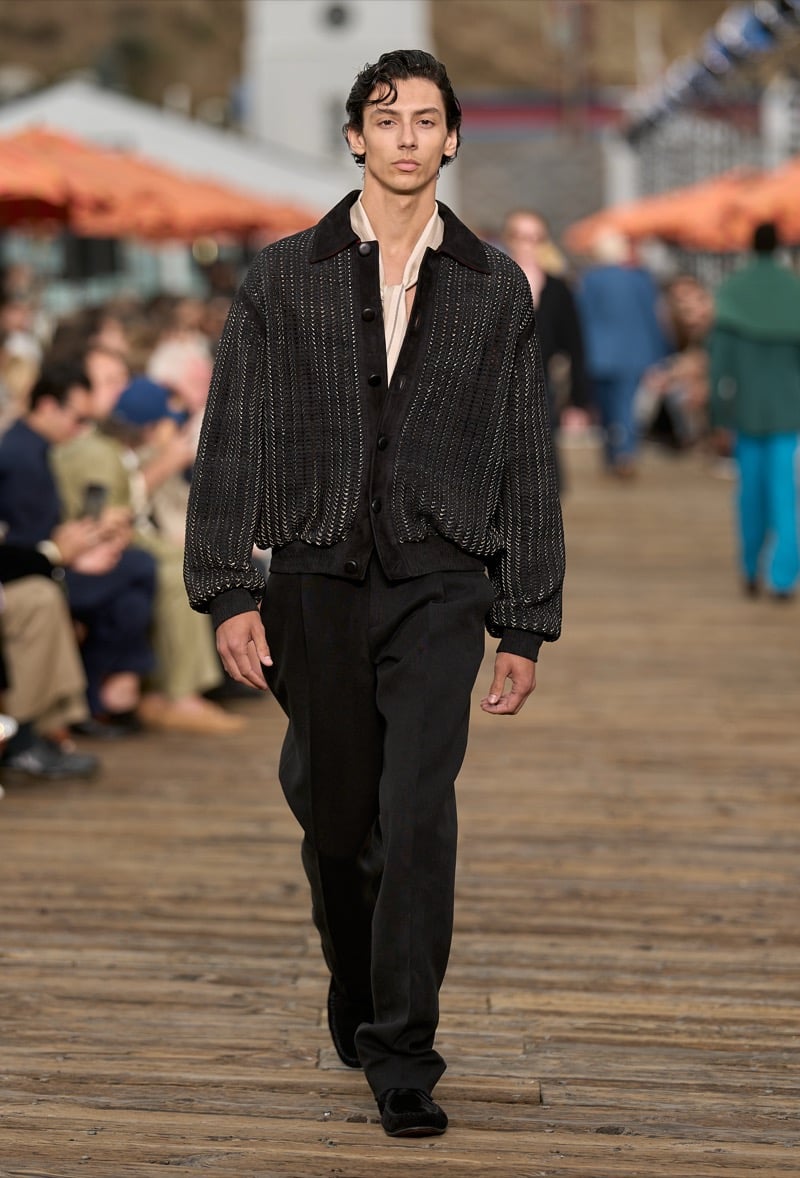 Male model wears a black beaded mesh knit bomber jacket over a blush silk shirt with charcoal pleated wide-leg trousers and black velvet shoes