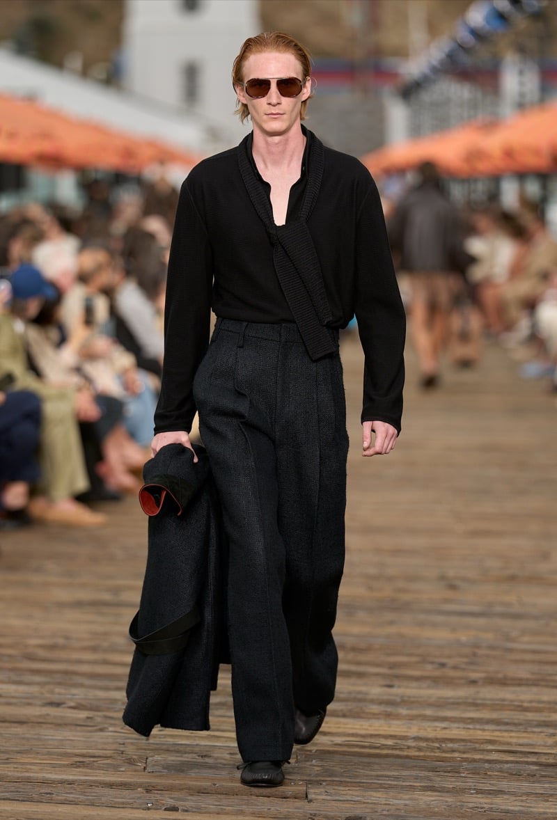 Male model wears a black ribbed knit henley with a knit sash collar over high-waisted dark wide-leg trousers, carrying a folded black coat with rust lining