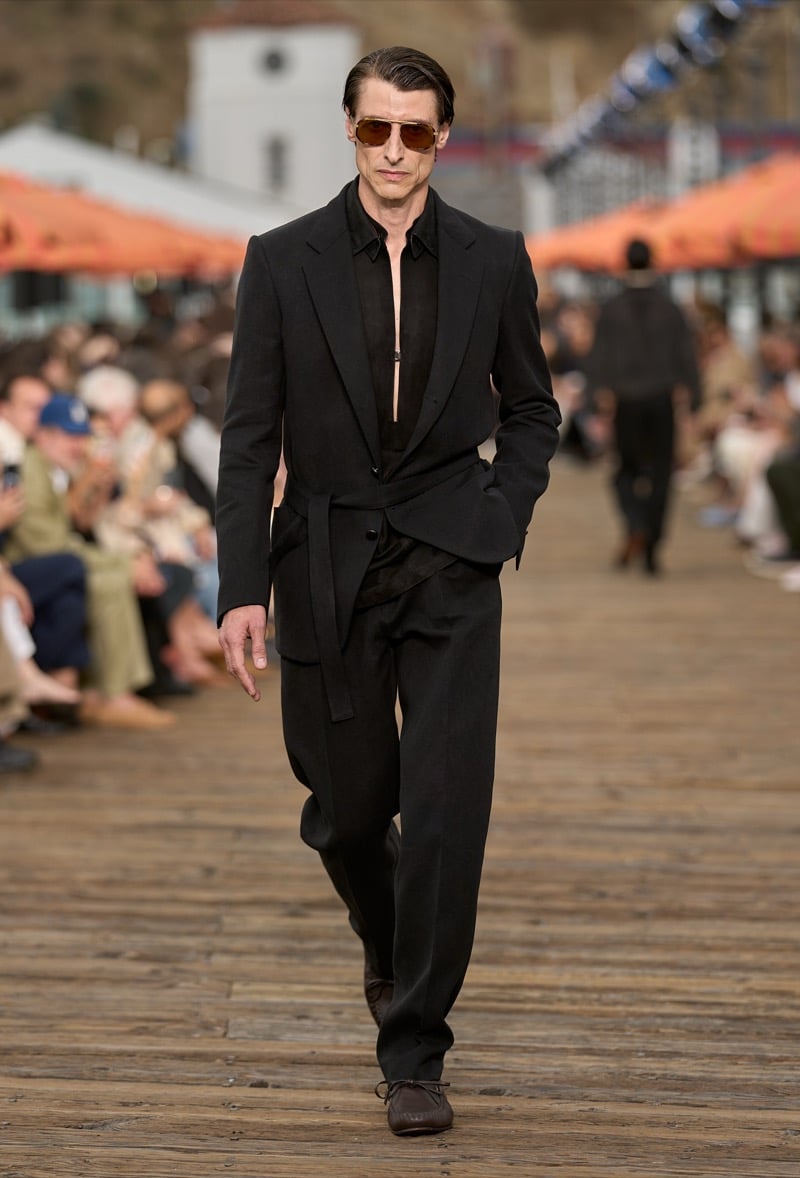Male model wears a black peak lapel belted single-breasted suit over an open black silk shirt with brown lace-up moccasins and tortoiseshell aviator sunglasses