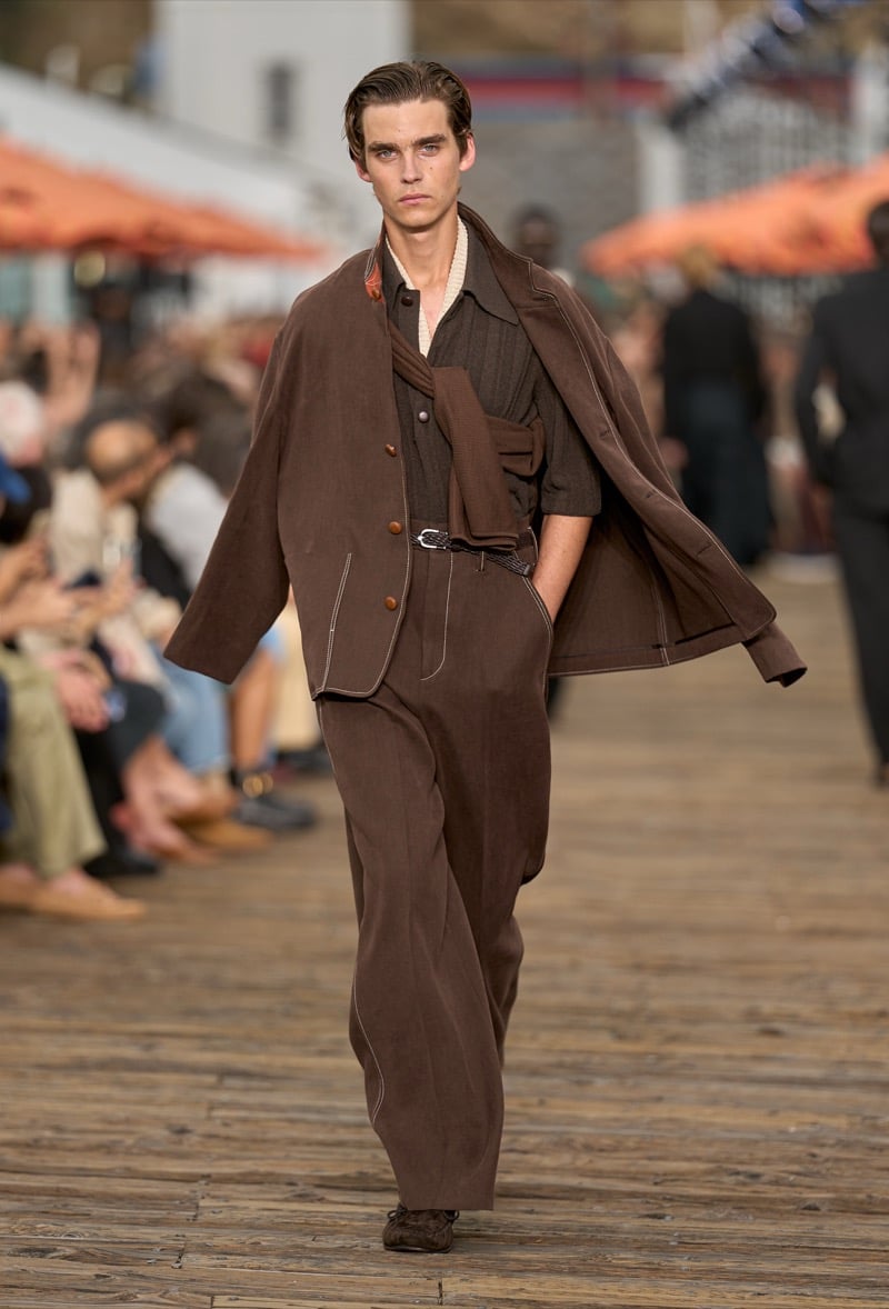 Male model wears a chocolate brown belted blazer with contrast stitching over a brown ribbed knit short-sleeve polo and matching wide-leg trousers, with a brown overcoat draped at the shoulders