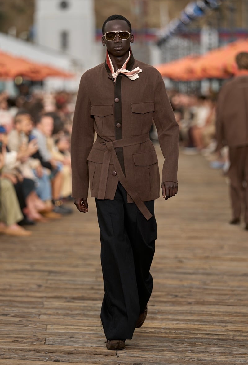 Male model wears a dark brown belted safari jacket over a black shirt with a blush silk scarf knotted at the neck, paired with black wide-leg trousers and brown moccasins