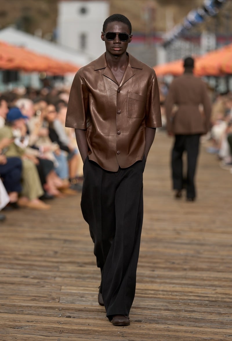 Male model wears a chocolate brown nappa leather short-sleeve camp shirt over black wide-leg trousers with brown leather pointed moccasins and black aviator sunglasses