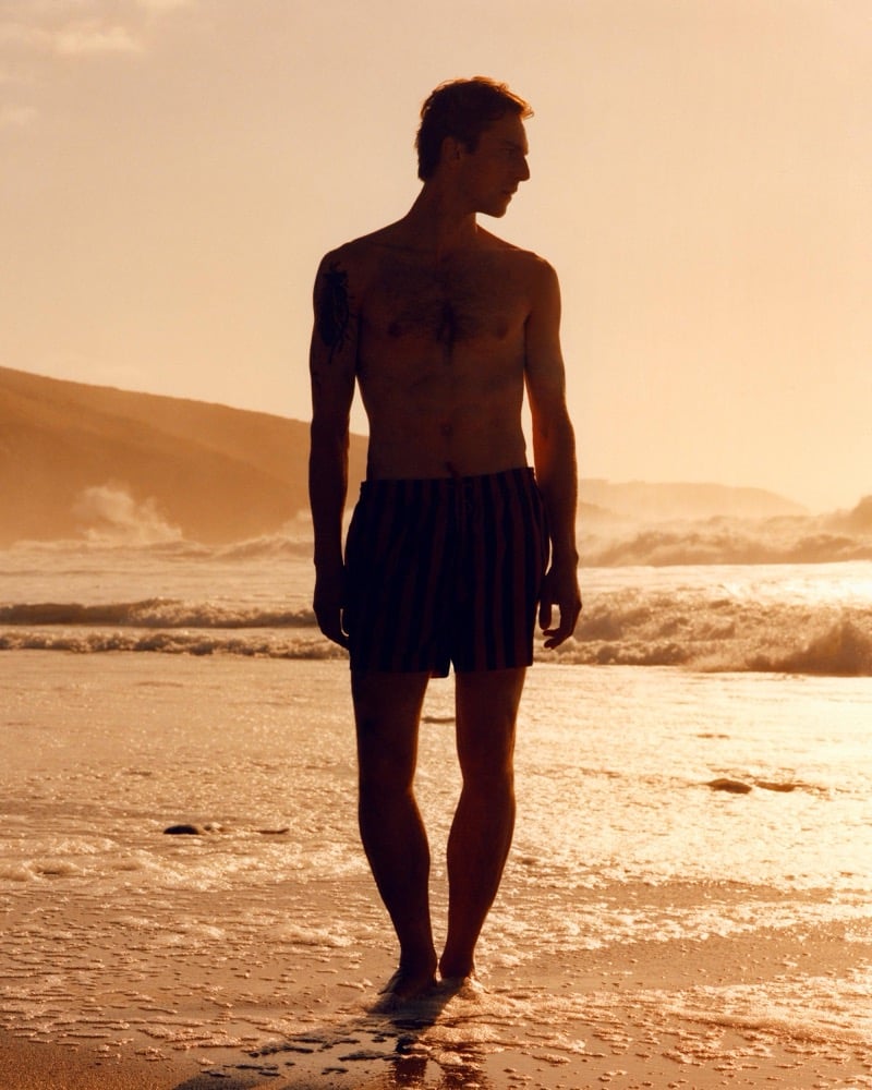 Silhouette of Quentin Demeester shirtless in striped swim shorts walking along the shoreline at sunset