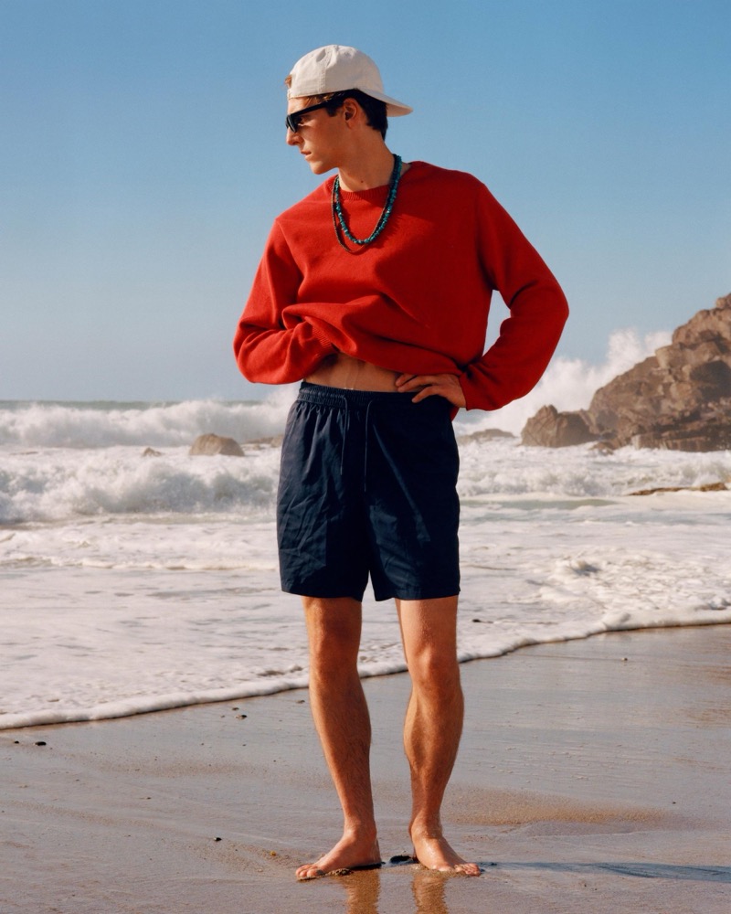 Quentin Demeester in a red crewneck sweater and swim shorts stands on the beach in a cap, sunglasses, and a turquoise beaded necklace as waves break behind him