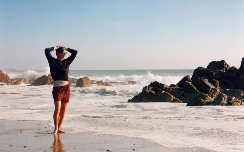 Quentin Demeester adjusts his cap on the beach in striped swim shorts and a cropped sweater, with rocky outcrops and crashing waves behind him