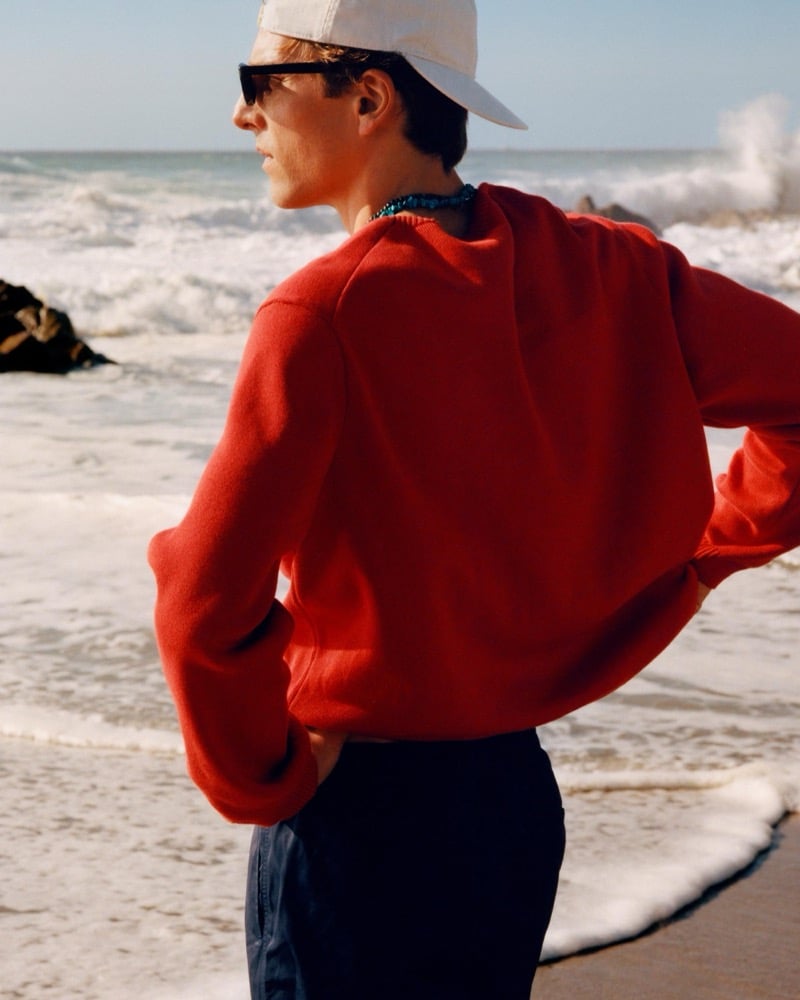 Quentin Demeester seen from behind in a red sweater and cap looking out at crashing waves on the beach
