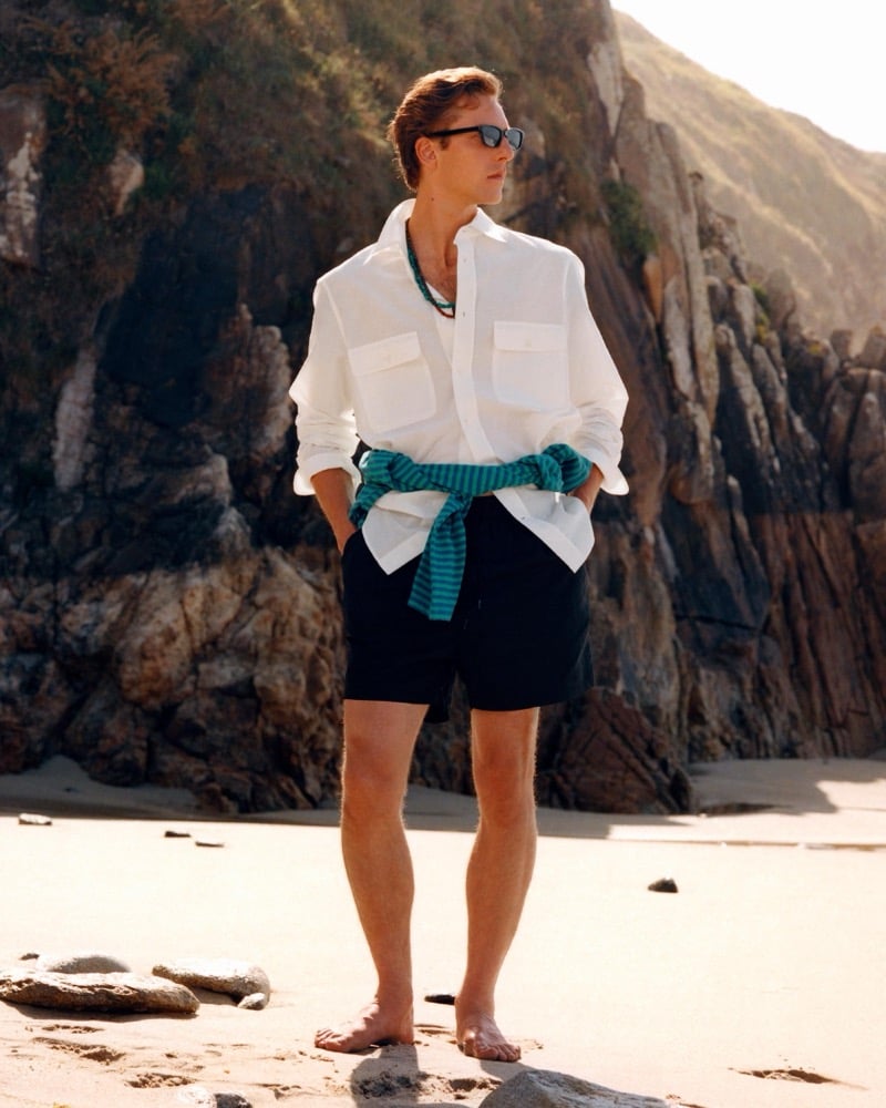 Quentin Demeester in a white utility shirt with a striped teal top knotted at the waist stands before a rocky cliff in swim shorts