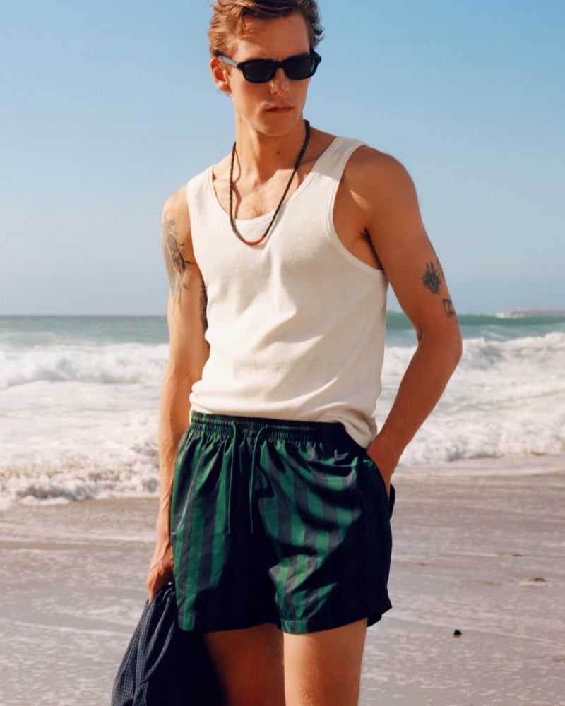Quentin Demeester in a white tank top and green striped swim shorts stands on the beach holding a folded garment, tattoos visible on both arms