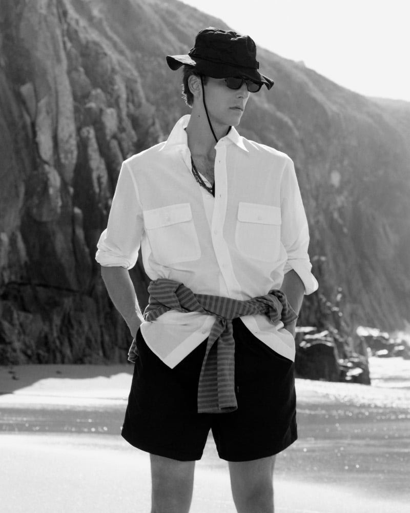 Black-and-white photo of Quentin Demeester in a bucket hat and white shirt with a striped top tied at the waist standing before a rocky cliff