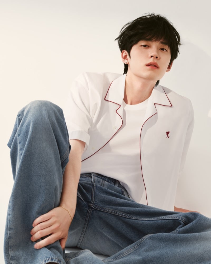 Zhou Yiran sits in a white camp-collar shirt trimmed with burgundy piping over a white T-shirt with the Ami heart logo, paired with baggy denim jeans, for Ami Paris' Sweetheart capsule