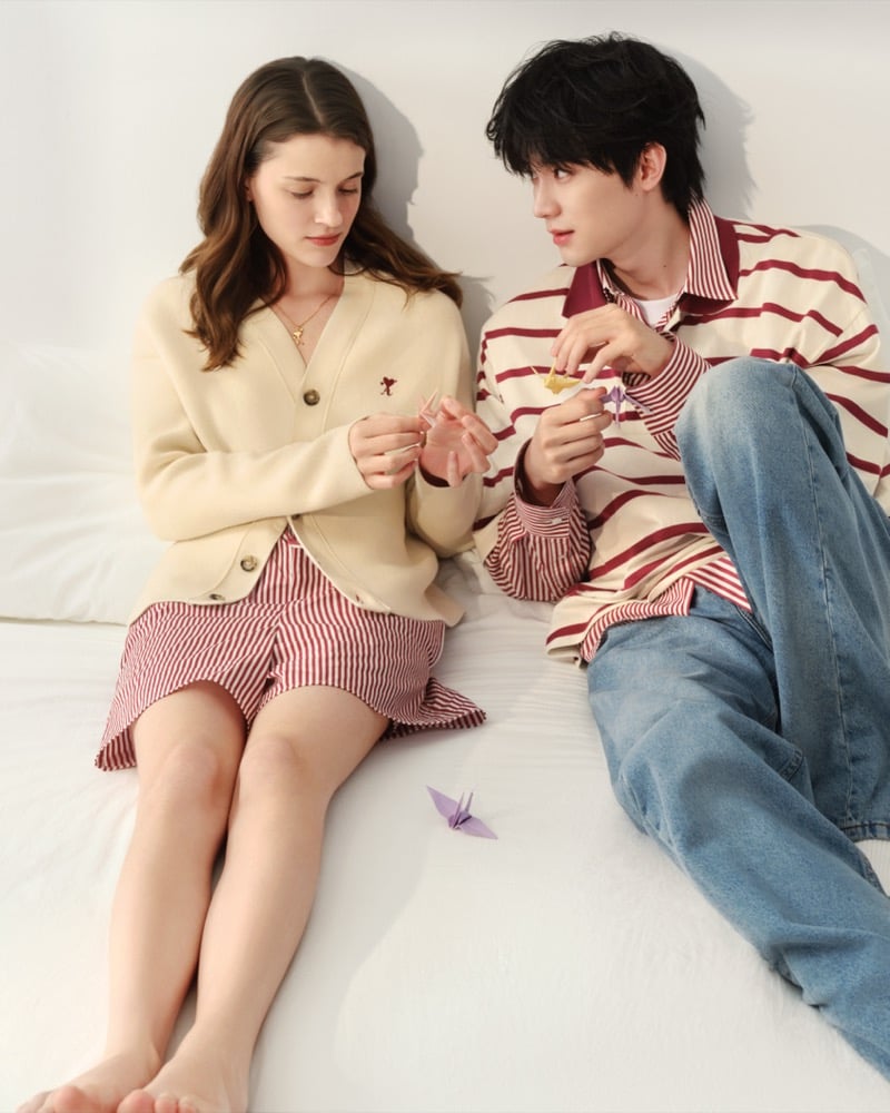 Zhou Yiran and a model sit side by side on a bed folding a paper crane, he in a striped polo and jeans, she in a cream cardigan and striped shorts, for Ami Paris' Sweetheart capsule