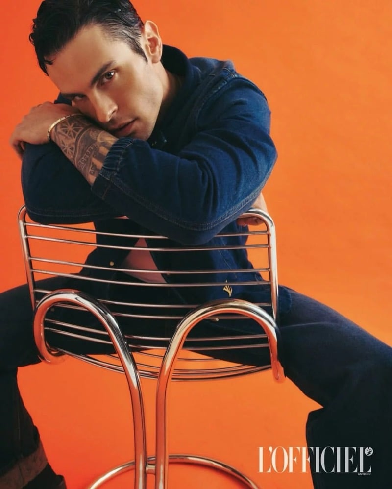 Baptiste Giabiconi rests his tattooed arm on a chrome chair back, wearing a dark wash denim jacket against an orange backdrop