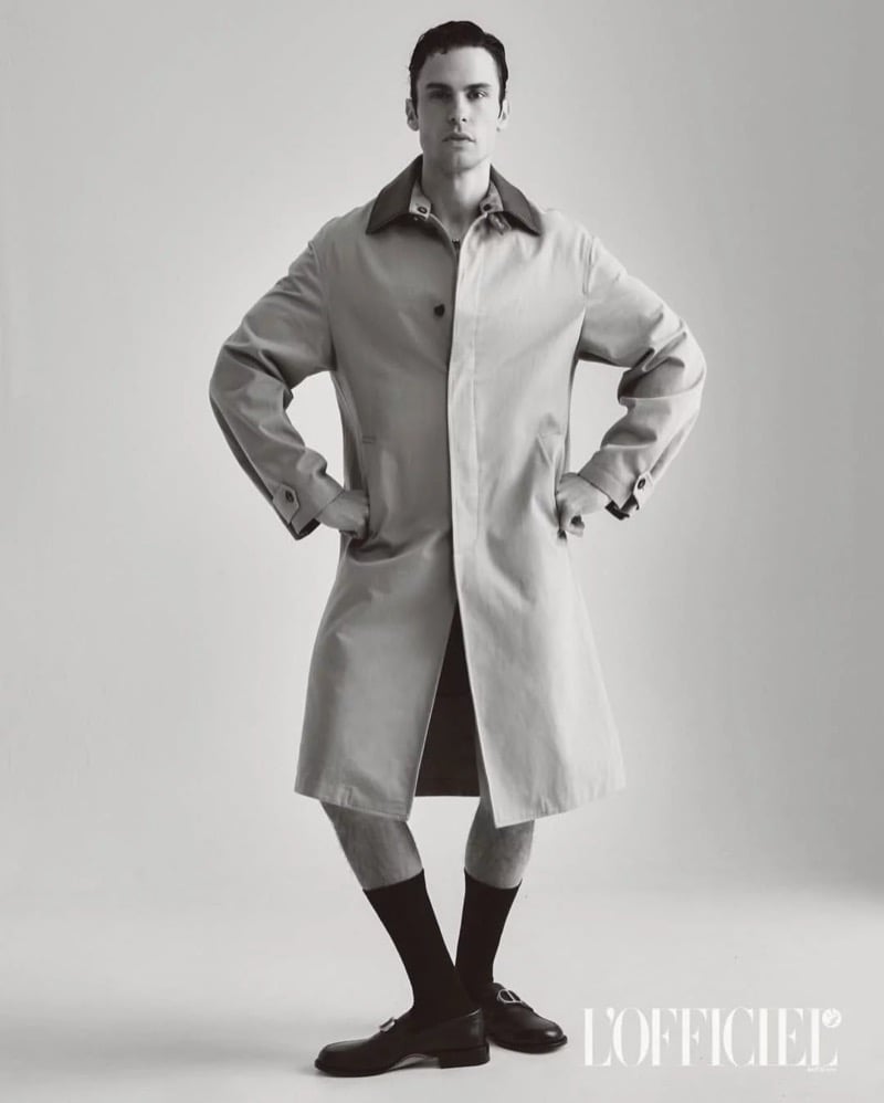 Baptiste Giabiconi stands in an oversized trench coat with buckled loafers against a plain studio backdrop