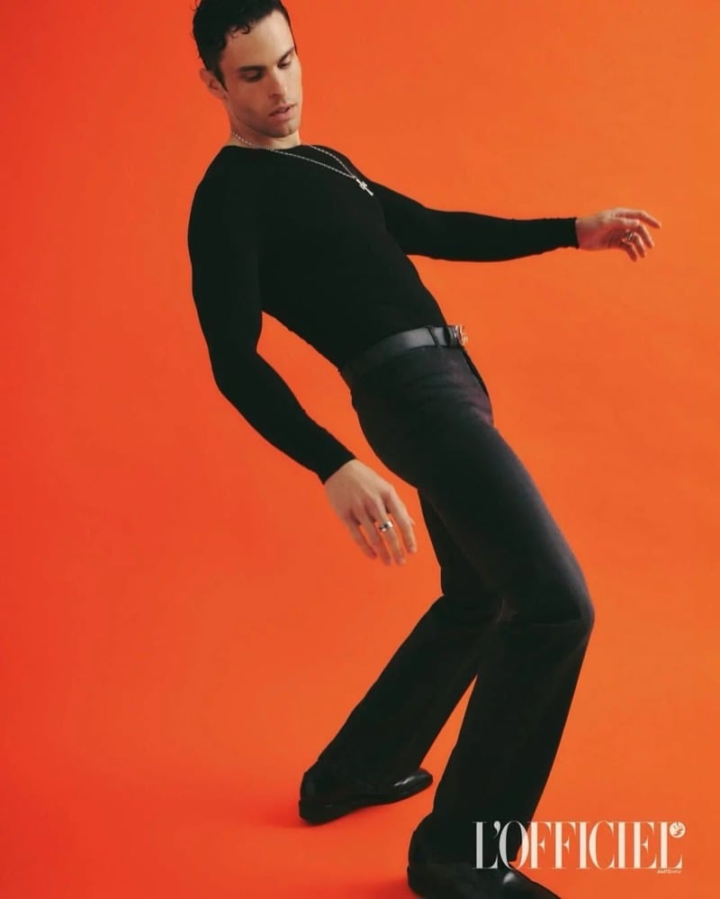 Baptiste Giabiconi leans into a dance-like pose in a black long-sleeve top and flared trousers against an orange backdrop
