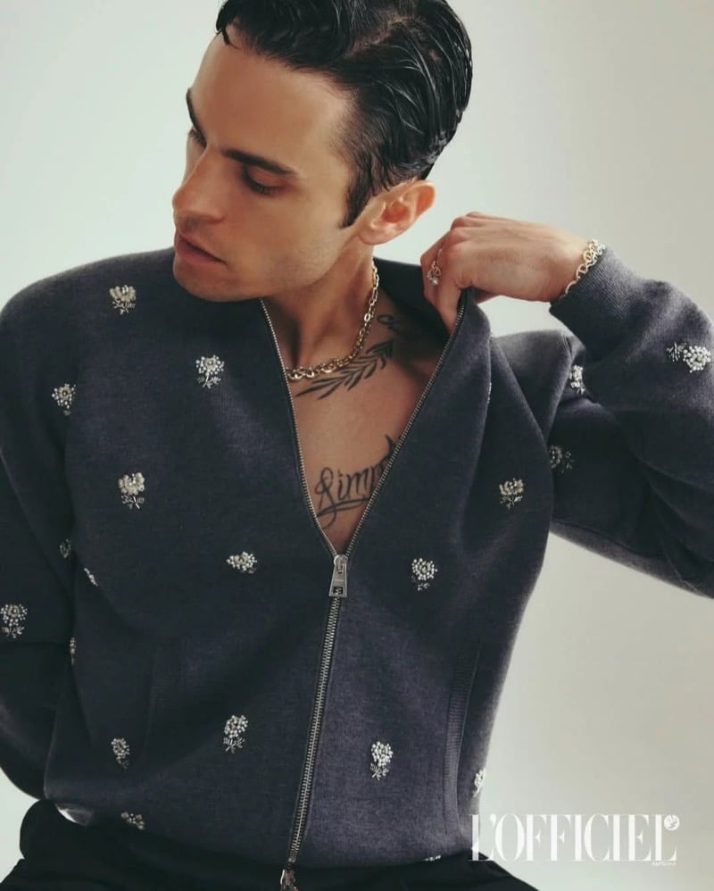 Baptiste Giabiconi adjusts the collar of a gray floral-embroidered Givenchy cardigan, chain necklace and chest tattoos visible