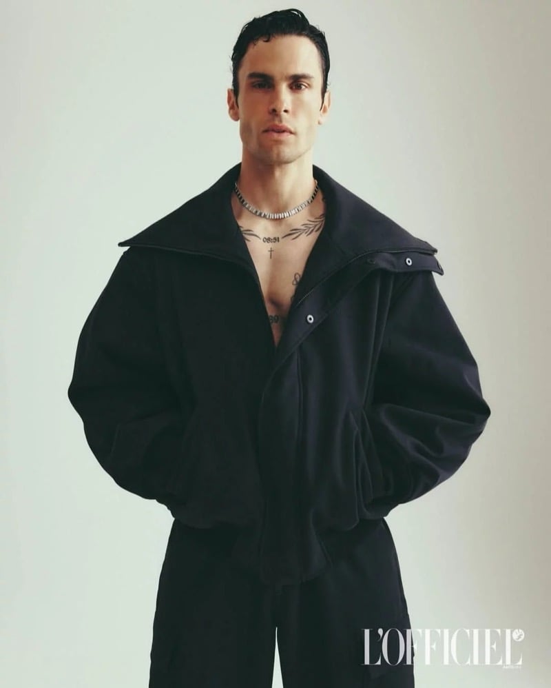 Baptiste Giabiconi wears an oversized black jacket with a high collar over his bare chest and tattoos