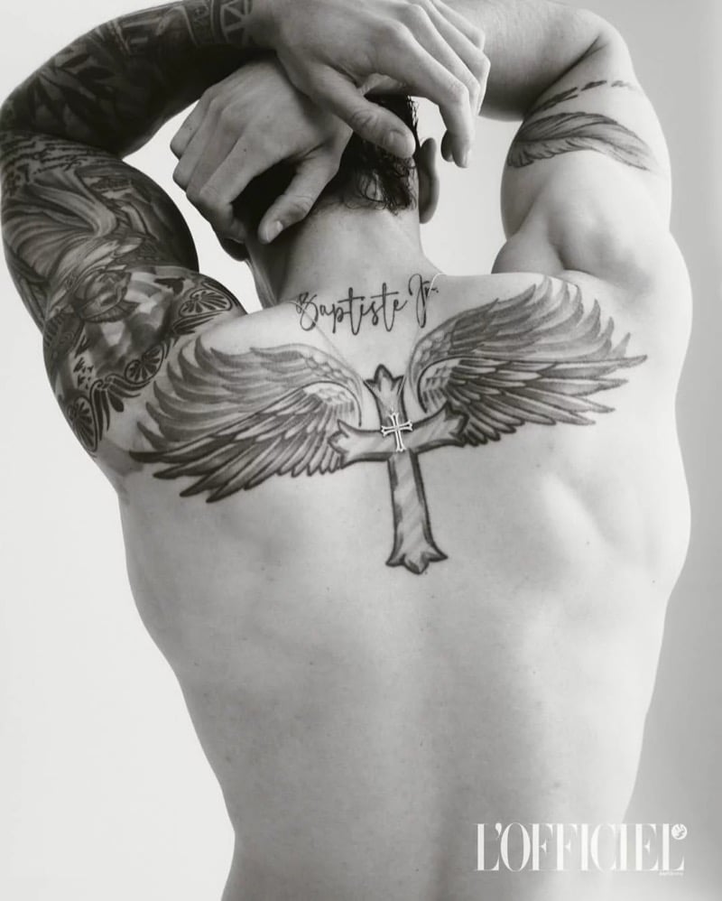 A black-and-white close-up of Baptiste Giabiconi's back tattoo of angel wings and a cross, arms raised behind his head