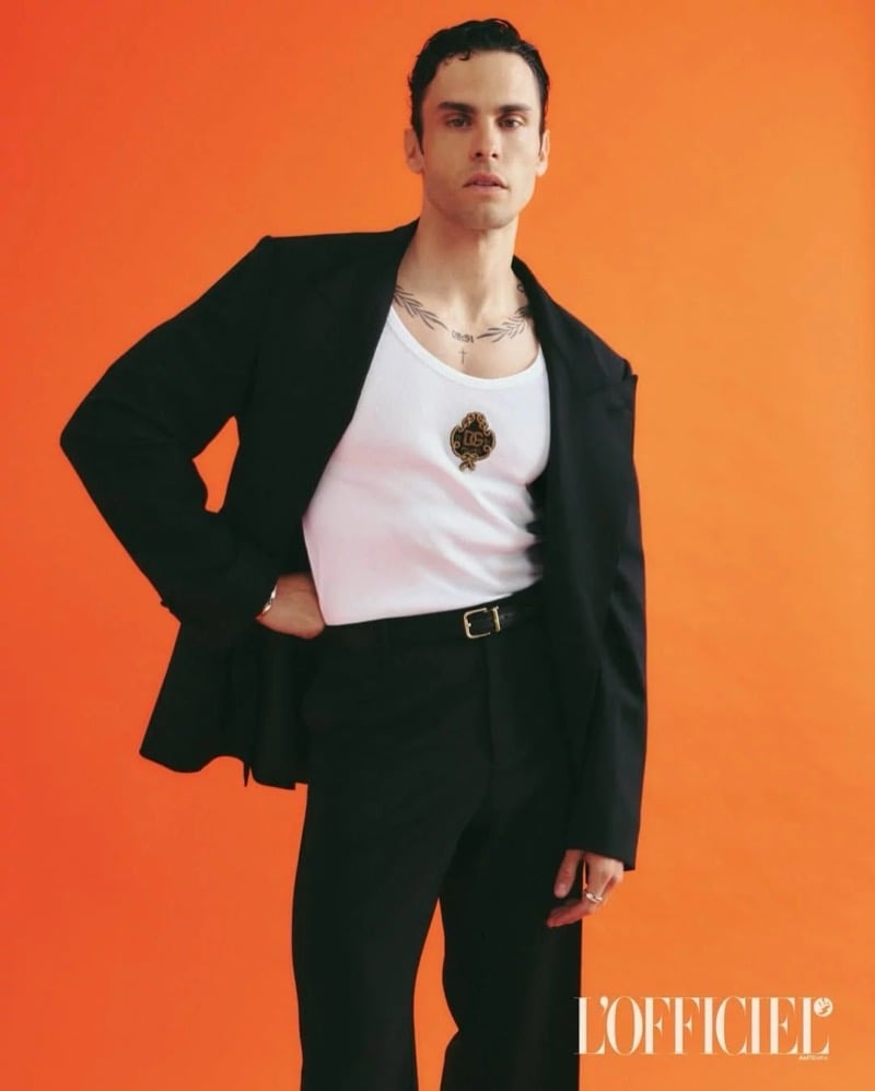 Baptiste Giabiconi wears an open black suit jacket over a white Dolce & Gabbana tank top against an orange backdrop