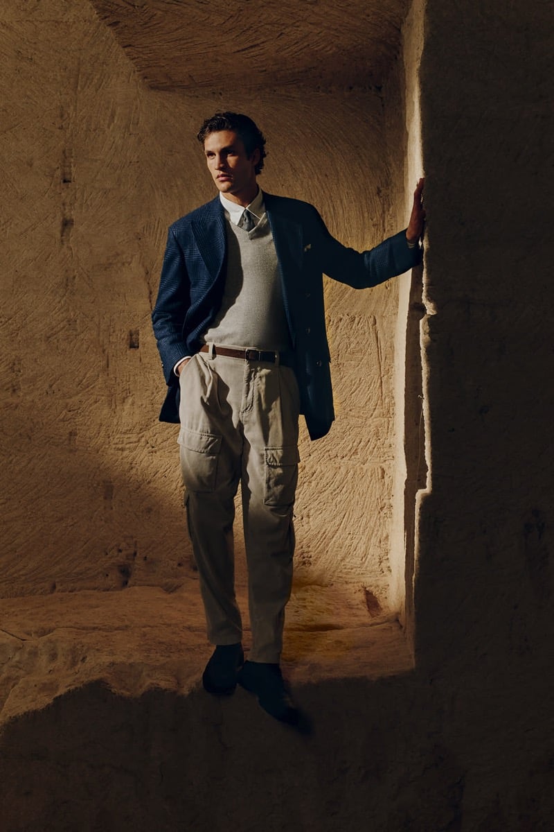 Francisco Henriques wears a navy blazer over a gray V-neck knit and white shirt with pleated khaki cargo trousers, standing in a sunlit stone passageway for Brunello Cucinelli's fall 2026 edit