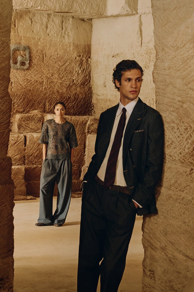 Francisco Henriques wears a double-breasted suit with a burgundy tie in a stone passageway, a fellow model in an embellished top standing out of focus behind him, for Brunello Cucinelli's fall 2026 edit