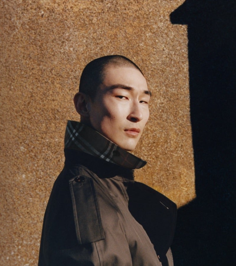 Profile portrait of Sang Woo Kim in Burberry's trench coat with a check collar against a sunlit textured wall