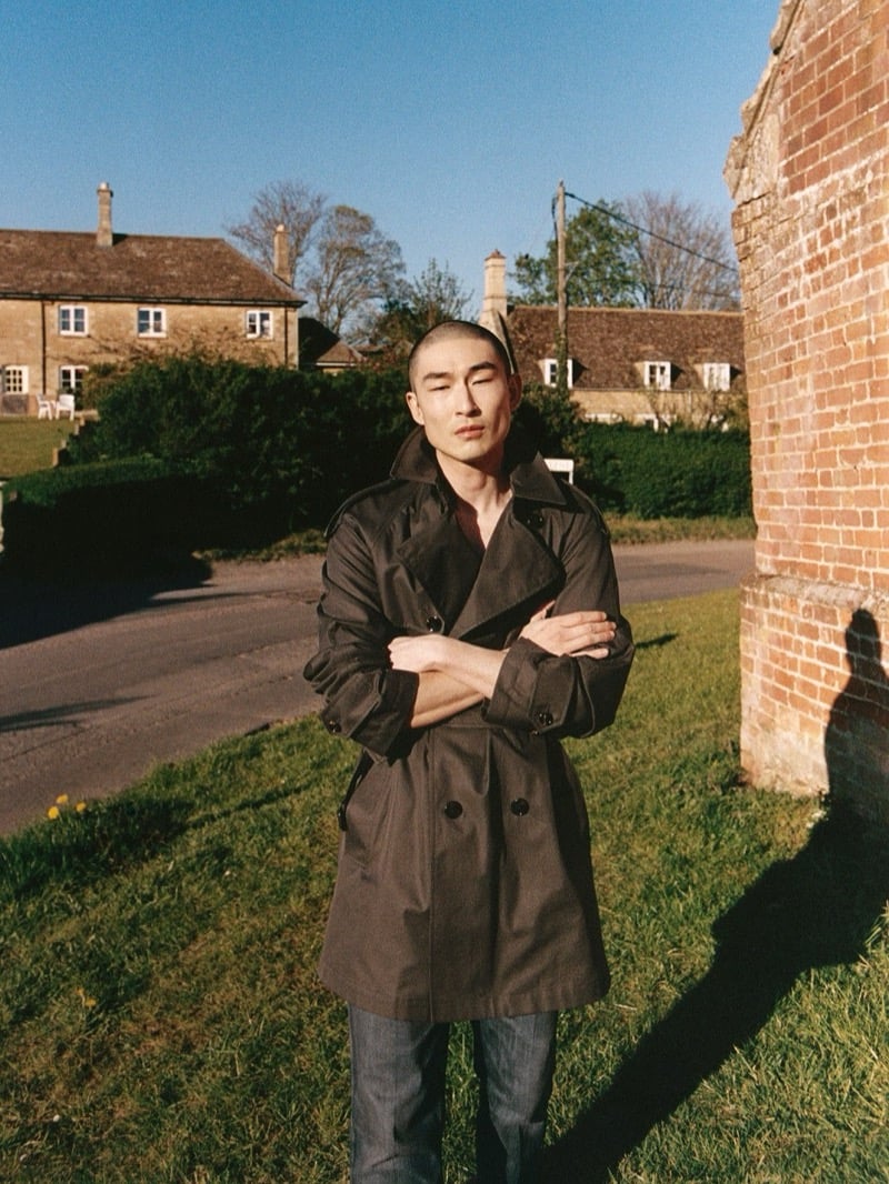 Sang Woo Kim crosses his arms in Burberry's Leadenham trench coat beside a brick wall in a Northamptonshire village