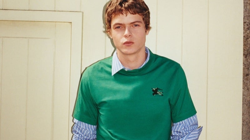 Moses Martin wears Burberry's green EKD logo T-shirt layered over a striped shirt against a paneled door