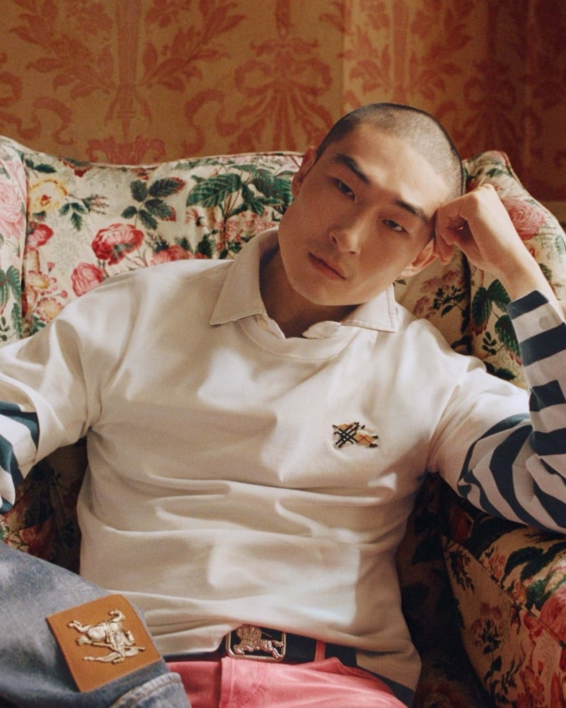 Sang Woo Kim reclines on a floral armchair wearing Burberry's EKD logo t-shirt layered over a striped top