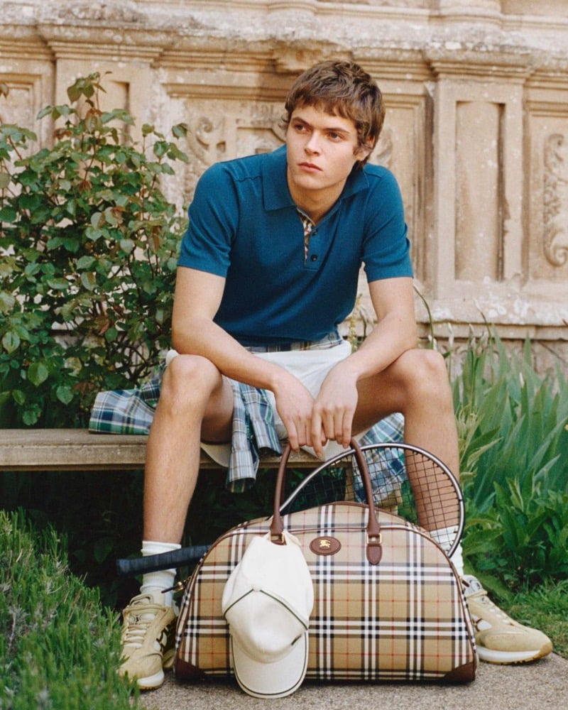 Moses Martin sits on a stone bench in a teal Burberry polo shirt and white shorts, a tennis racket resting against the brand's Vintage Check holdall