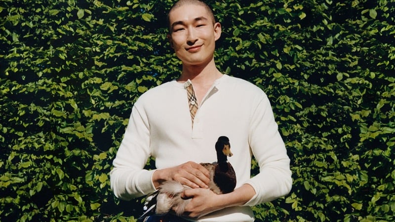 Sang Woo Kim smiles while holding a duck, wearing Burberry's henley with a check placket in front of a trimmed hedge
