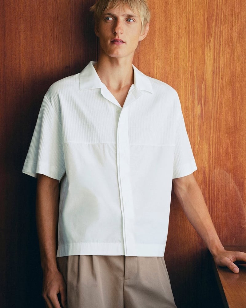 Pierrick Grégoire in a white ribbed-paneled camp shirt and tan trousers against a wood-paneled wall