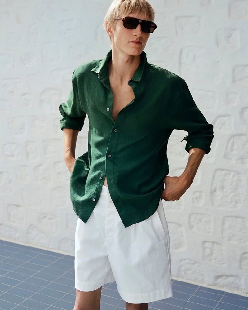 Pierrick Grégoire in a half open forest green linen shirt and white shorts with sunglasses against a textured white wall
