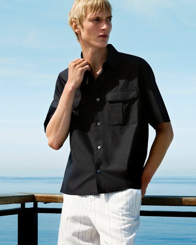 Pierrick Grégoire in a black camp shirt with a chest pocket and pinstriped trousers against the sea