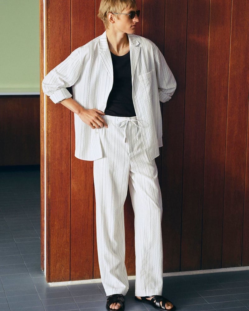 Pierrick Grégoire in a white pinstriped suit over a black T-shirt with black sandals against a wood-paneled wall