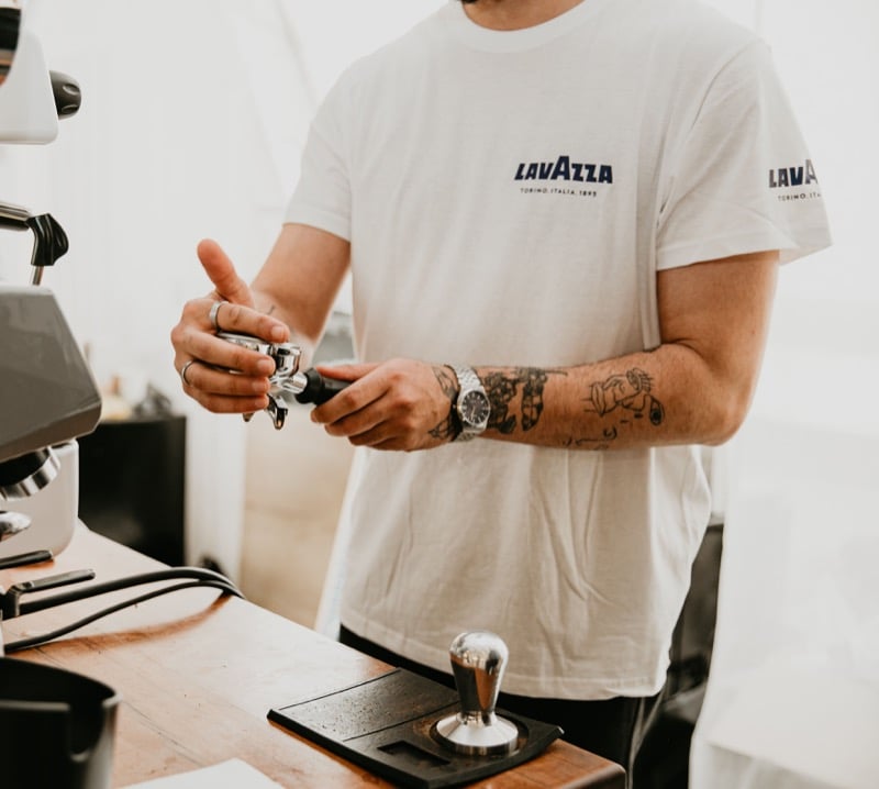A barista in a white company-branded t-shirt operates an espresso machine portafilter