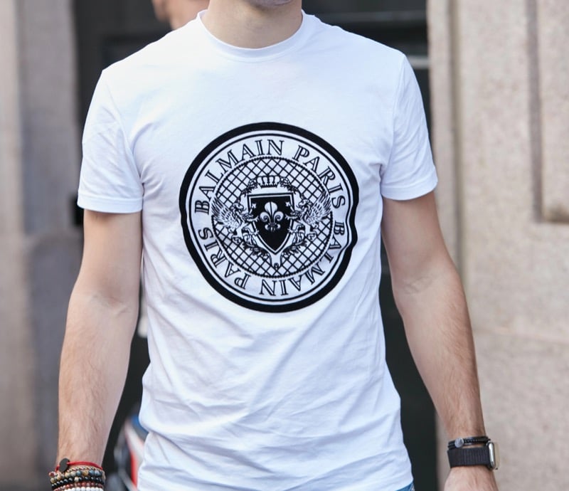 A close-up of a man wearing a white t-shirt with a black crest logo printed across the chest