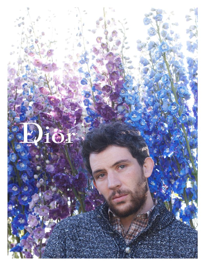 Josh O'Connor in a sequined knit sweater over a plaid shirt stands before towering purple and blue delphinium stalks