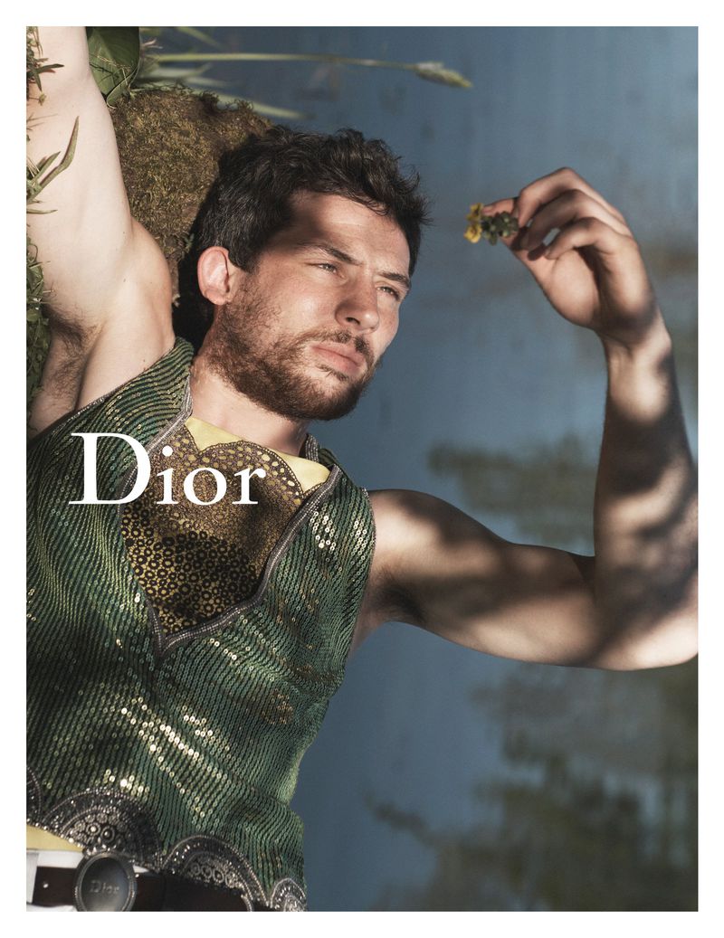 Josh O'Connor in a green and gold sequined top holds a small yellow flower overhead