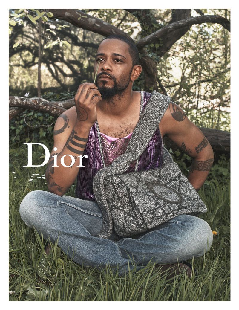 LaKeith Stanfield sits cross-legged in grass in a purple sequined tank top, a gray Dior bag on his shoulder, a stem raised to his lips