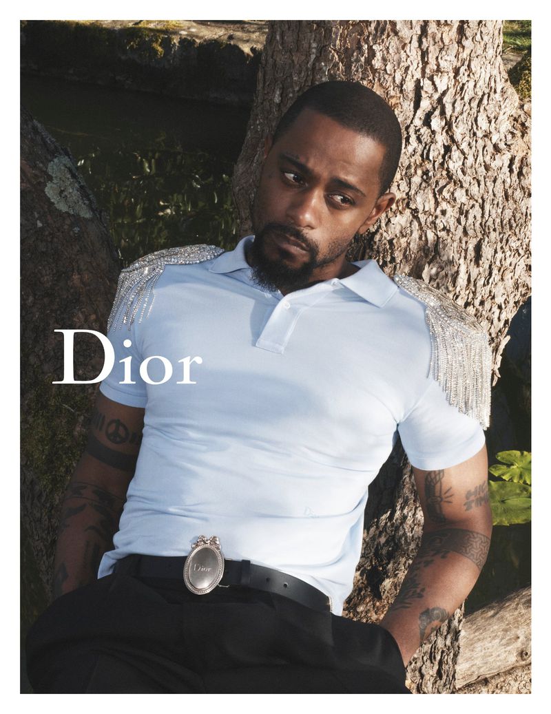 LaKeith Stanfield stands against a tree trunk by the water in a pale blue polo with a crystal-fringed shoulder, tattoos visible on both arms, a Dior belt buckle at his waist