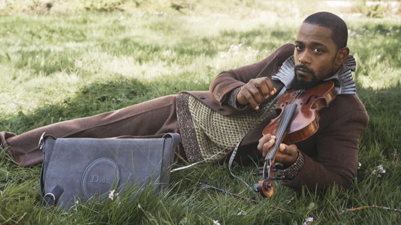 LaKeith Stanfield reclines in the grass playing a violin, wearing a brown wool coat with a ruffled collar beside a Dior suede bag
