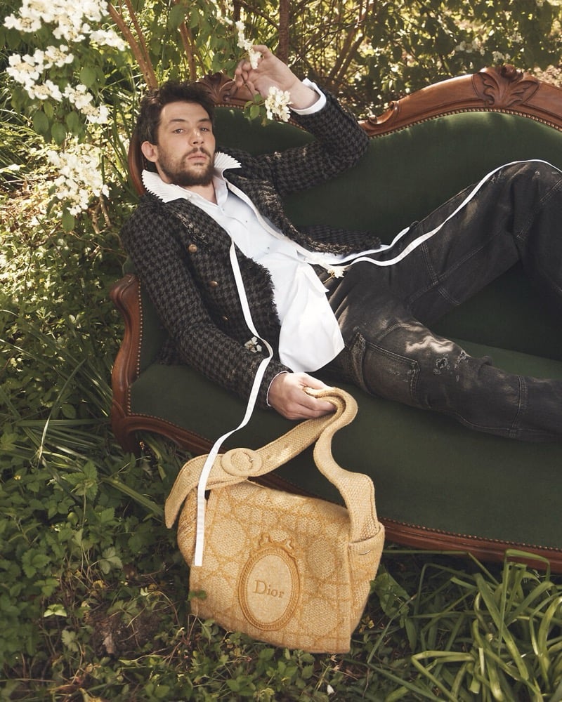 Josh O'Connor reclines on an antique green velvet sofa beneath flowering branches, in a houndstooth jacket with a ruffled collar, holding a woven Dior tote