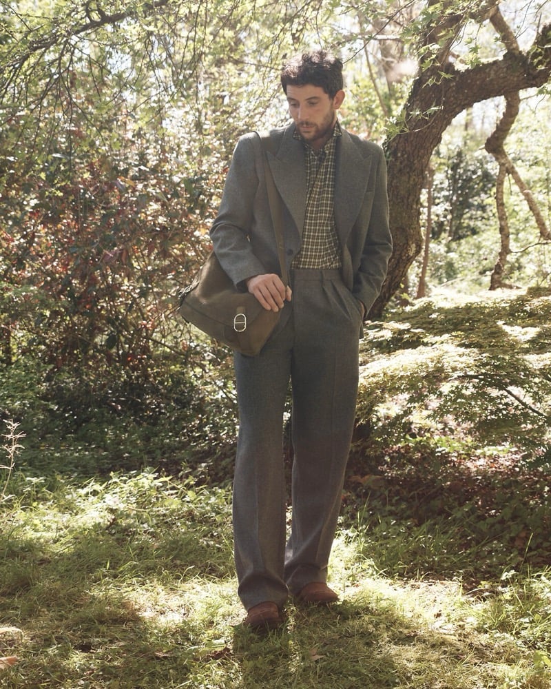 Josh O'Connor stands in a wooded clearing in a gray wool suit and checked shirt, a brown leather Dior bag on his shoulder