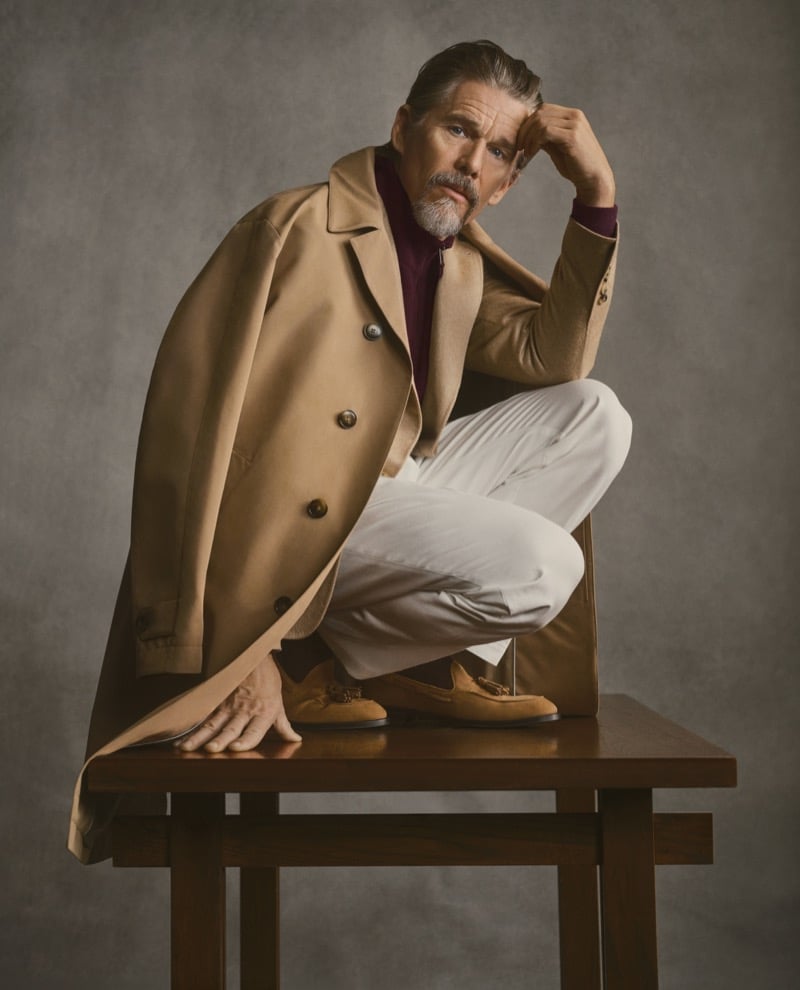Ethan Hawke crouches on a wooden stool in a camel double-breasted coat over a burgundy zip-neck sweater with tasseled suede loafers, for Canali's fall-winter 2026 campaign